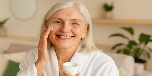 A smiling senior woman in a white robe applies face cream while holding a jar, sitting in a softly lit room with green plants.